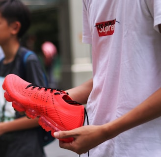 A person holding a vibrant orange sports shoe with translucent soles, standing next to someone else. The person is wearing a white t-shirt with a distinct logo in red and black. The background is muted, suggesting an urban environment.