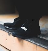 A close-up view of black athletic shoes with a visible brand logo, placed on a concrete step. The shoes are stylishly designed and appear to be suitable for sports or casual wear. The background is blurred, focusing attention on the footwear.