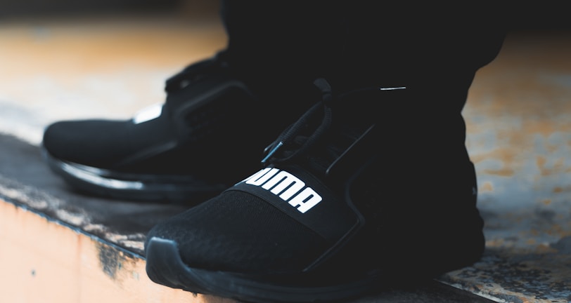 A close-up view of black athletic shoes with a visible brand logo, placed on a concrete step. The shoes are stylishly designed and appear to be suitable for sports or casual wear. The background is blurred, focusing attention on the footwear.
