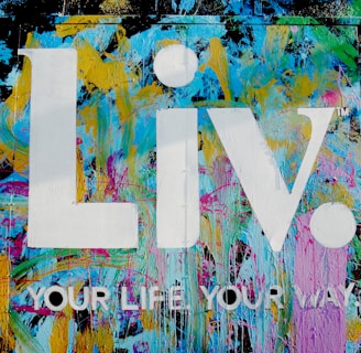 The image features a vibrant graffiti-style background with splashes of blue, yellow, pink, and green colors. Superimposed on this colorful backdrop are the large white letters 'Liv.' and the phrase 'YOUR LIFE YOUR WAY.' in additional white text.
