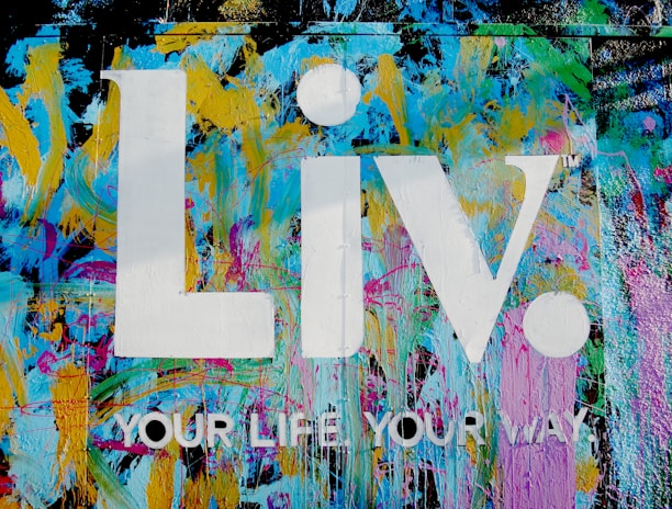 The image features a vibrant graffiti-style background with splashes of blue, yellow, pink, and green colors. Superimposed on this colorful backdrop are the large white letters 'Liv.' and the phrase 'YOUR LIFE YOUR WAY.' in additional white text.