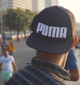 A person wearing a cap with the brand name 'Puma' stands on a bustling waterfront promenade. Various people are walking or standing around, with tall buildings and greenery lining the background. The ocean is visible to the right, adding to the relaxed yet busy atmosphere.