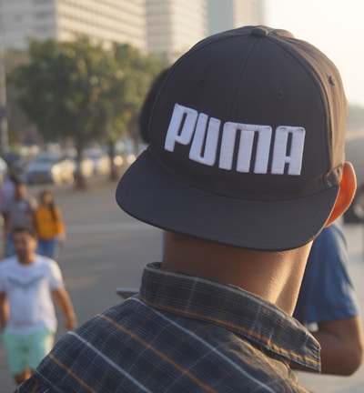 A person wearing a cap with the brand name 'Puma' stands on a bustling waterfront promenade. Various people are walking or standing around, with tall buildings and greenery lining the background. The ocean is visible to the right, adding to the relaxed yet busy atmosphere.