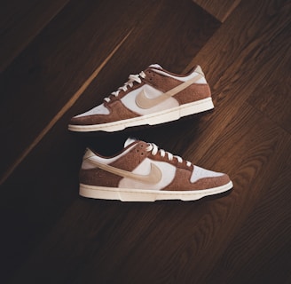 A pair of brown and white sneakers with prominent branding positioned on a dark wooden floor. The stylish shoes have a low-top design with white shoelaces and a distinctive swoosh logo on the sides.