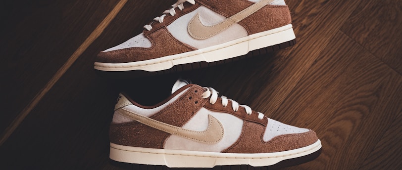 A pair of brown and white sneakers with prominent branding positioned on a dark wooden floor. The stylish shoes have a low-top design with white shoelaces and a distinctive swoosh logo on the sides.