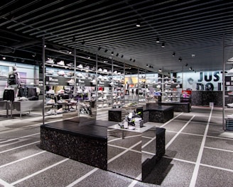 A modern sneaker store with sleek black shelving displaying various athletic shoes and sports apparel. Central seating areas and spotlights provide a contemporary retail ambiance. The signage prominently features a famous sportswear slogan in the background.