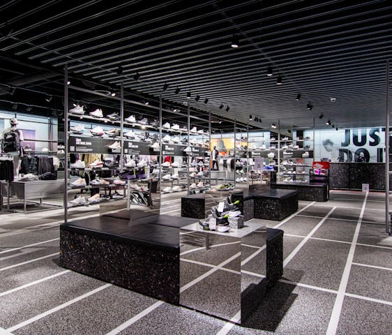 A modern sneaker store with sleek black shelving displaying various athletic shoes and sports apparel. Central seating areas and spotlights provide a contemporary retail ambiance. The signage prominently features a famous sportswear slogan in the background.