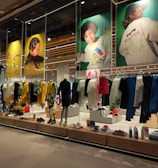 A retail store showcases clothing and sportswear on mannequins and hangers, with large vibrant posters featuring children in sports attire above. The interior displays bright garments and accessories on shelves and racks.