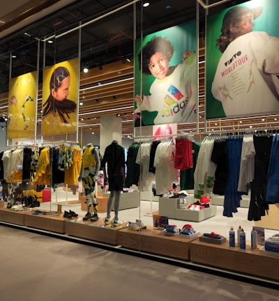 A retail store showcases clothing and sportswear on mannequins and hangers, with large vibrant posters featuring children in sports attire above. The interior displays bright garments and accessories on shelves and racks.