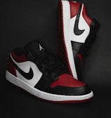 A pair of stylish athletic shoes in black, white, and red colors with distinctive branding. The shoes are positioned against a dark background, showcasing their sleek design and detailed craftsmanship.