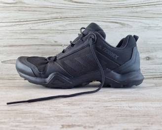 A black athletic shoe with a rugged sole and mesh detailing, designed for outdoor activities. It features brand markings on the side and is positioned against a light wooden background.