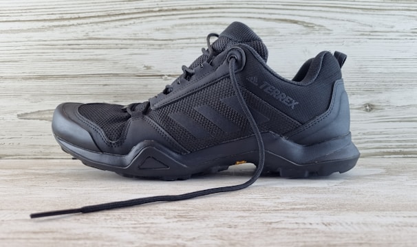 A black athletic shoe with a rugged sole and mesh detailing, designed for outdoor activities. It features brand markings on the side and is positioned against a light wooden background.