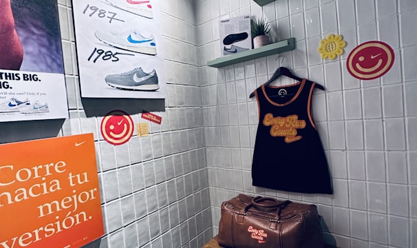 A small retro-themed display with a focus on athletic apparel and shoes. The walls feature a series of posters showcasing various Nike shoes from different years. A black gym tank top and a brown leather gym bag are prominently positioned on a wooden bench. There are decorative elements like potted plants on wall-mounted shelves and smiley face icons. The environment has a nostalgic and sporty atmosphere.